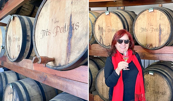 Deusa Rodrigues com óculos de sol em loja de barris de carvalho, segurando uma taça de vinho branco, nas prateleiras de cachaça Três Pedras. Rotas Gastronômicas do interior de SP.