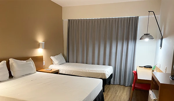 Quarto de hotel com duas camas, iluminação suave, cortina grande e mesa de trabalho com cadeira vermelha, ideal para hospedagem confortável.
