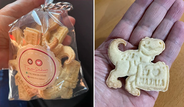Biscoito de Natal com personagem de dinossauro em forma de biscoito, embalado em saco transparente com selo personalizado, e no outro presenting um biscoito de dinossauro artesanal na mão.