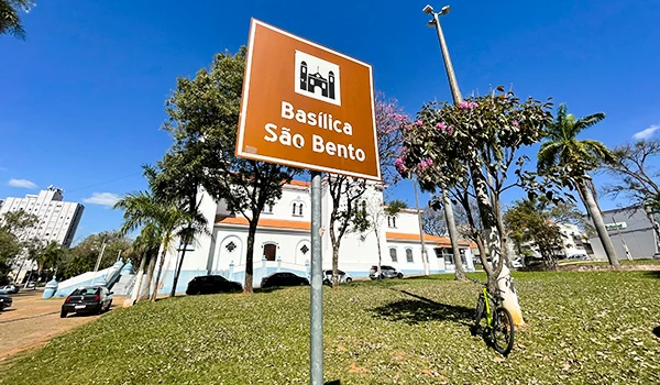 Sinalização indicando a direção para a Basílica São Bento localizada em uma área com árvores, carros e uma bicicleta estacionada ao lado, com céu azul ao fundo em Marília.