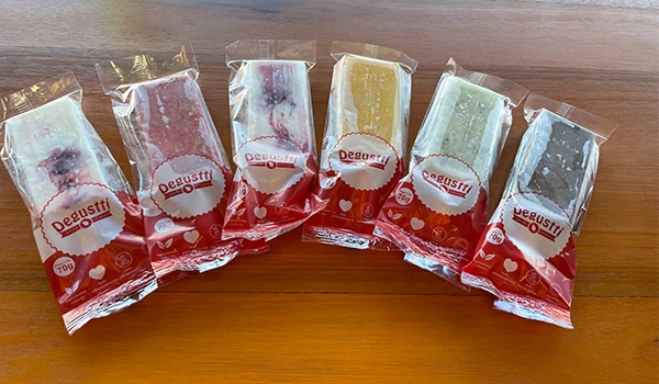 Imagem de diversos tipos picolés em diferentes cores, embalados da marca Degusti, organizados sobre uma mesa de madeira. Rotas Gastronômicas do interior de SP.