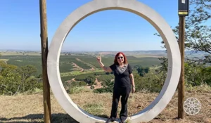 Deusa Rodrigues paz dentro de uma estrutura circular ao ar livre, com vista panorâmica de campos e vegetação, sob céu claro
