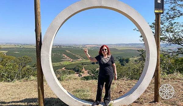 Deusa Rodrigues paz dentro de uma estrutura circular ao ar livre, com vista panorâmica de campos e vegetação, sob céu claro