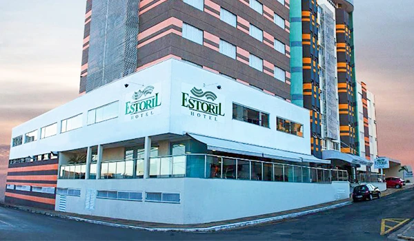 Imagem do Hotel Estoril, edifício moderno localizado em uma área urbana, com vários andares e fachada branca, refletindo um pôr-do-sol ao fundo.