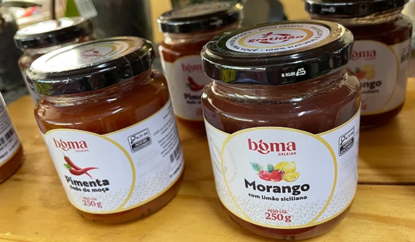 Diversos potes de geleia de morango com embalagem de 250g, produção da marca Boma, exibidos em uma loja ou feira de alimentos.