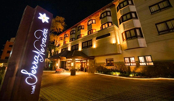 Fachada do Hotel Serra Nevada iluminada à noite, com sinalização destacada ao lado e ambiente acolhedor na entrada.
