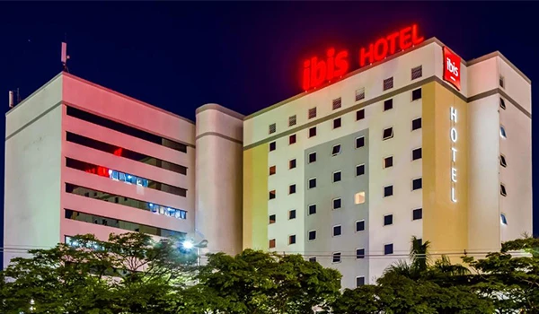 Imagem do ibis hotel à noite com letreiro luminoso e fachada moderna, rodeado por árvores e céu escuro ao fundo, destacando-se na paisagem urbana.
