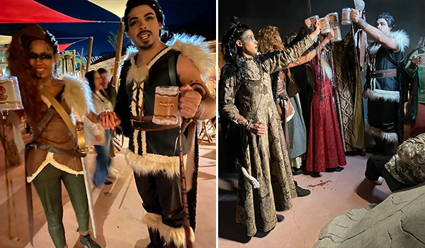 Imagem de pessoas curtindo uma festa tradicional, com pessoas vestidas com roupas típicas e brindando com cerveja, ambiente animado com luzes e barracas.