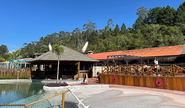 Imagem de um espaço ao ar livre com área de piscina, estrutura de madeira e área de lazer sob céu azul claro, cercada por vegetação e árvores. Vila Viking e o “Quarto do Rei”.