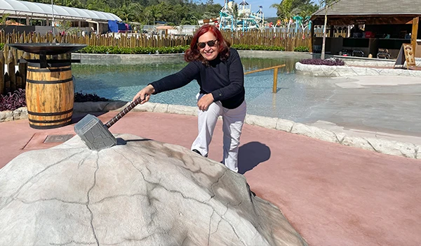 Deusa Rodrigues brincando na Vila Vijing segurando um martelo do Thor em uma estrutura de rocha artificial, com fundo de lago e brinquedos aquáticos ao fundo.