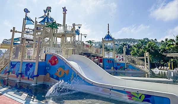 Parque aquático com escorregador e área de diversão infantil decorada com elementos marinhos, em um dia ensolarado com céu parcialmente nublado.