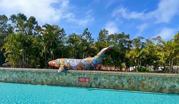 Escultura de lagarto colorido na borda de uma piscina, com vegetação ao fundo e céu azul claro, em ambiente externo tropical.