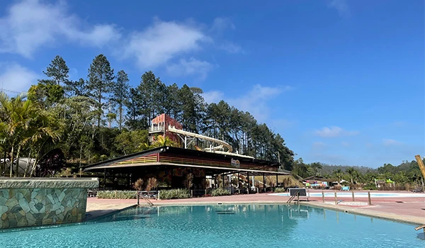 Piscina ao ar livre com área de lazer em um hotel ou resort situado em meio à natureza, com árvores e céu azul ao fundo, ideal para descanso e lazer.