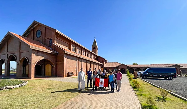 Grupo de pessoas em frente ao Mosteiro da Divina Misericórdia em Maríla, com arquitetura tradicional, com fachada de tijolos e estacionamento ao lado, sob céu claro.