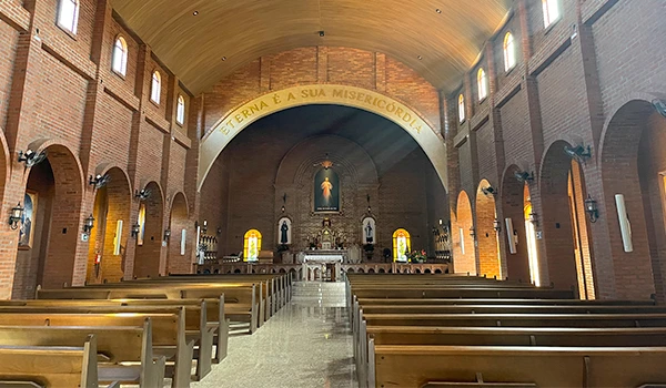 Interior de uma igreja moderna com bancos de madeira, altares e janelas coloridas, destacando a mensagem "Eterna é a sua misericórdia" no arco central.