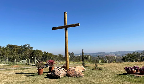 Cruz de madeira ao ar livre em um campo com céu azul, cercado por plantas e uma cerca de arame, simbolizando fé e espiritualidade.