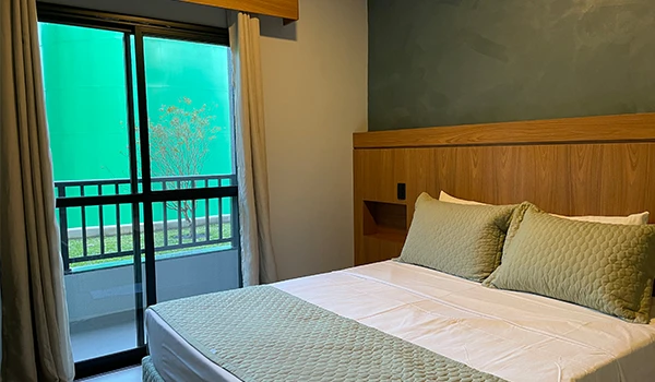 Quarto de hotel elegante com cama confortável, almofadas decorativas, parede verde acentuada e varanda com vista para o jardim, criando um ambiente acolhedor.