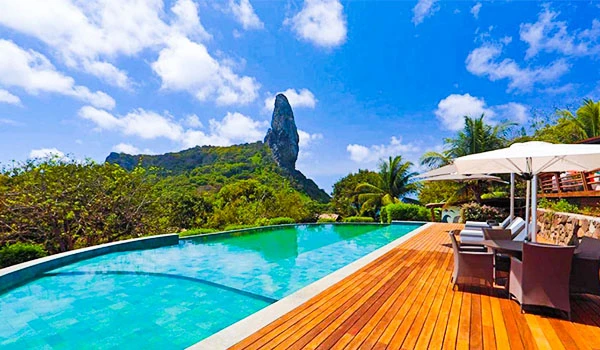 Piscina ao ar livre com vista para natureza exuberante e montanha em fundo, área de lazer com espreguiçadeiras e guarda-sol, ambiente relaxante e perfeito para descanso.