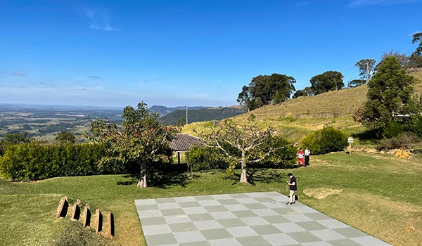 Cenário ao ar livre com uma área de xadrez gigante, árvores, pessoas jogando e vista para as colinas e céu azul, ideal para lazer e momentos de diversão ao ar livre.