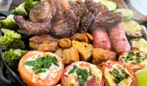 Prato com variedade de carnes e acompanhamentos, incluindo brócolis, tomates recheados com queijo, e batatas assadas, preparado para uma refeição saborosa.Rotas Gastronômicas no Interior SP.
