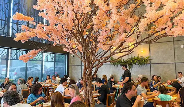 Imagem do Restaurante Don Mantelli de Marília com decoração de árvore de flores de cerejeira artificial no centro, rodeada por clientes desfrutando de suas refeições e bebidas, criando um ambiente aconchegante e elegante.