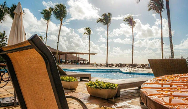 Imagem mostrando uma área de piscina ao ar livre com espreguiçadeiras, palmeiras e um ambiente de resort ensolarado. Um lugares para comemorar Bodas de Prata no Brasil.