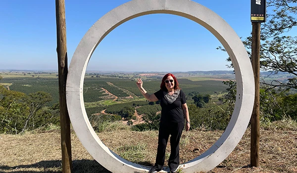 Deusa Rodrigues posando ao lado de uma estrutura circular em um campo aberto com vista para o horizonte, usando roupas pretas e óculos de sol.