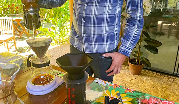 Pessoa preparando café de forma artesanal com método pour-over, usando filtro de papel em uma cafeteria ao ar livre, com plantas ao fundo. Rotas Gastronômicas do interior de SP.