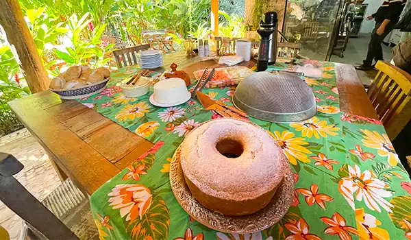 Mesa de jantar decorada com toalha floral, tendo um bolo de rosquinha em destaque, rodeada de comida, utensílios e ambientes ao ar livre com plantas.