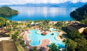 Vibrante vista de resort com piscina de formato irregular, rodeada por palmeiras, com vista para um lago ou mar, em uma paisagem tropical exuberante.