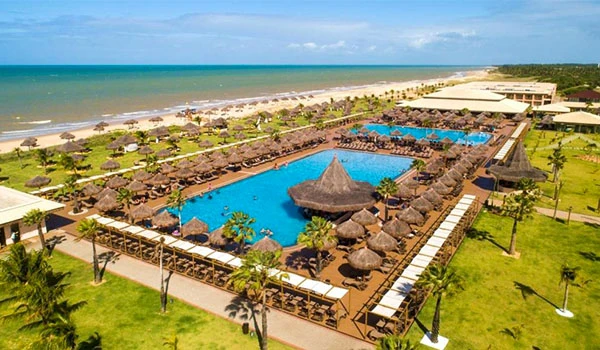 Vista panorâmica de um resort à beira-mar com piscina gigante ao centro, muitos chapéus de palha, área de lazer, e praia de areia branca ao fundo, ideal para relaxar.
