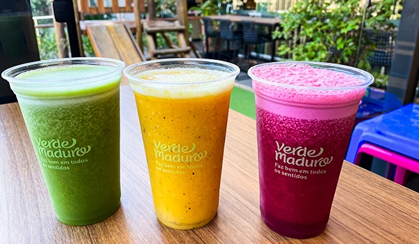 Três sucos coloridos alinhados sobre uma mesa de madeira na varanda de um café ou restaurante ao ar livre, com várias plantas e móveis ao fundo, promovendo uma atmosfera relaxante. Rotas Gastronômicas do interior de SP.