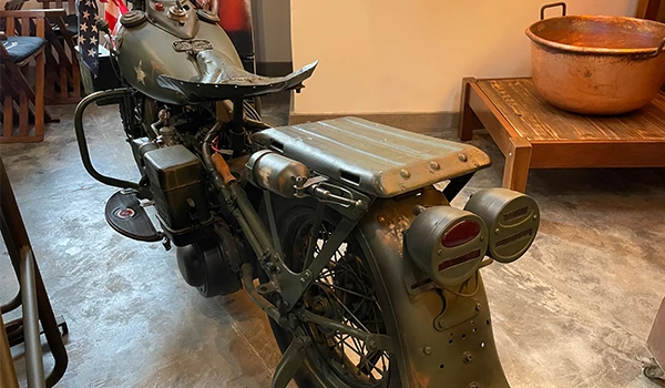 Imagem de uma motocicleta vintage em estilo militar, com acabamento envelhecido, mostrada em ambiente interno com piso de cimento e móveis de madeira ao fundo.