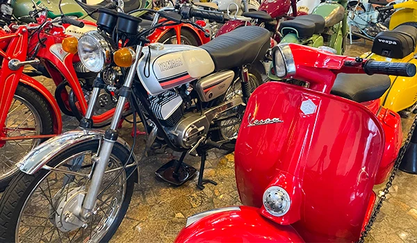 Coleção de motos clássicas e scooters vintage, incluindo uma Vespa vermelha e motocicletas de época, exibidas em um ambiente de museu. 