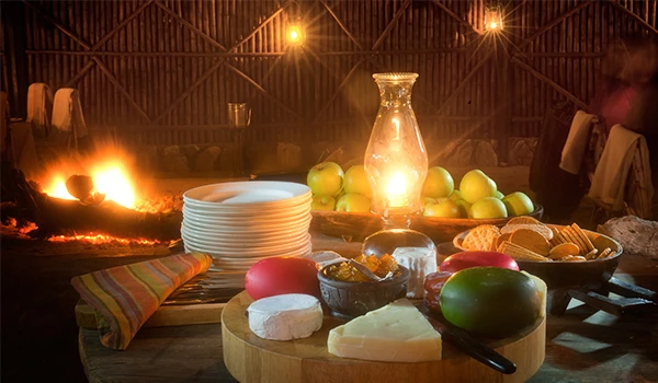 Mesa posta em uma decoração rústica com frutas, queijos, pães e uma vela de vidro em um ambiente acolhedor com iluminação quente. Dicas do Boma jantar savanas África.