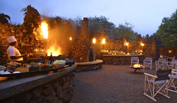 Cena de um ambiente ao ar livre ao entardecer com fogueiras, cadeiras de madeira, uma churrasqueira e decoração rústica, ideal para eventos ao ar livre. Dicas do Boma jantar savanas África.