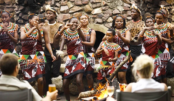 Grupo de pessoas afro-brasileiras dançando e celebrando ao redor de uma fogueira, usando roupas tradicionais coloridas em cerimônia cultural. Dicas do Boma jantar savanas África.