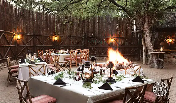 Decoração para jantar ao ar livre em ambiente rústico, com mesas elegantemente decoradas, velas e uma fogueira ao fundo, perfeito para eventos ao ar livre.