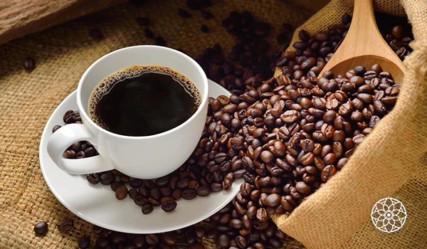 Imagem de uma xícara de café preto sobre um prato branco, ao lado de grãos de café em uma sacola de pano, destacando o aroma e sabor do café.