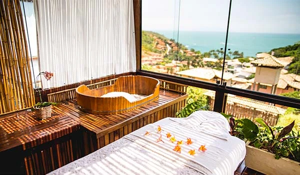Sala de massagem com vista panorâmica para a cidade e o oceano, incluindo banheira de hidromassagem, penteadeira de madeira e plantas decorativas. Armação dos Búzios hotéis Lua de Mel.