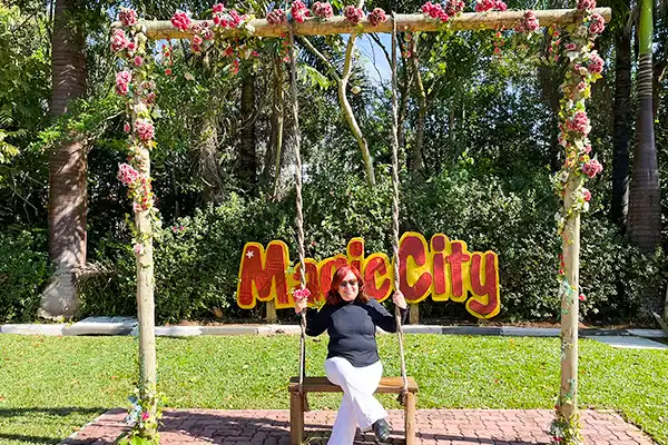 Mulher sentada em um balanço decorado com flores no parque Mooca City, com fundo de árvores e grama, desfrutando de um dia ensolarado.