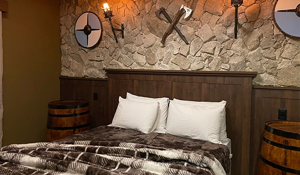 Imagem de uma cama de casal com cabeceira de madeira, travesseiros brancos e um edredom de pelúcia em um quarto aconchegante com decoração rústica na parede de pedra. Elementos decorativos em ferro na parede e lamparinas criam um ambiente acolhedor e sofisticado.