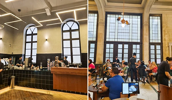 Foto de um café elegante com teto alto, janelas grandes e pessoas desfrutando de suas refeições e bebidas em ambiente aconchegante e bem decorado.