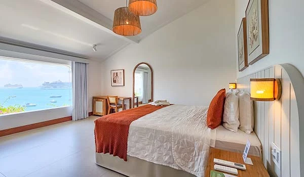 Quarto de hotel com vista para o mar, cama com roupa de cama branca, almofadas laranja, iluminação aconchegante e decoração moderna. Armação dos Búzios hotéis Lua de Mel.
