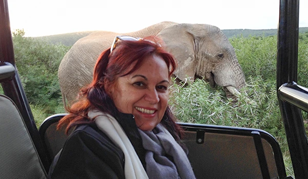 Deusa Rodrigues em safári, observando um elefante ao fundo, com natureza ao redor, explorando vida selvagem. Dicas do Boma jantar savanas África.
