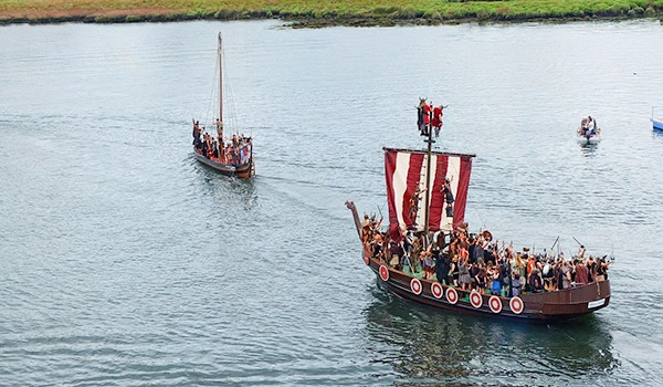Recriação histórica em barcos de época com pessoas fantasiadas, navegando em rio ou lago. Cenário ao ar livre, ideal para eventos culturais e turísticos.