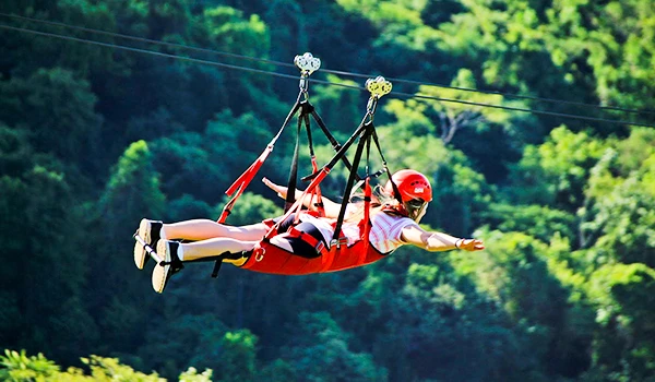 Pessoa fazendo tirolesa em meio à natureza, com árvores verdes ao fundo e usando equipamento de segurança, incluindo capacete vermelho.