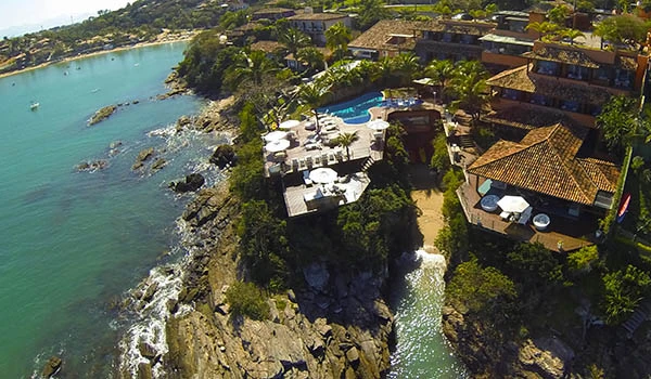 Vista aérea de hospedagem de luxo à beira-mar com piscina, rodeada por vegetação e casas, em frente ao oceano em um cenário paradisíaco. Armação dos Búzios hotéis para Lua de Mel.