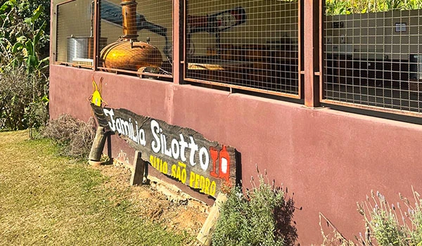 Imagem de fachada de Sitio São Pedro com anúncio que diz 'Família Silotto', localizado em Serra Negra, apresentando uma parede rosa clara, plantas ao redor e uma grade de proteção na parte superior.