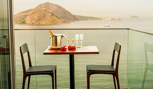 Varanda com vista para o mar, mesa decorada com flores, champanhe e balde de gelo, ideal para momentos românticos ou comemorações à beira-mar.